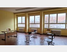 Leerer Klassenraum mit gelb gestrichenen Wänden, großen Fensterfronten und Heizkörpern darunter. Im Raum stehen vereinzelt Schulstühle und Tische auf einem braunen Boden; durch die Fenster sind Bäume und Dächer zu sehen.