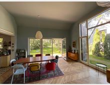 Esszimmer mit einem großen Holztisch und bunten Stühlen auf einem Teppich, helle Holzböden und eine Glasfront, die den Blick in einen Garten mit viel Grün freigibt. Rechts steht eine Holzkommode, links ist eine offene Küche erkennbar.