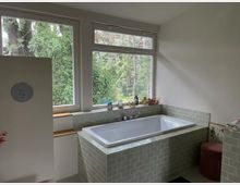 Helles Badezimmer mit eingebauter Badewanne, die von hellgrünen Kacheln umgeben ist. Große Fenster mit Blick auf eine bewaldete Umgebung lassen viel natürliches Licht herein, daneben befinden sich ein kleines Regal und Dekorationsgegenstände.