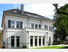 Zweistöckiges weißes Einfamilienhaus mit dunkelgrauem Walmdach und Fenstern mit Rollläden, umgeben von einem Garten mit Rasen und Bäumen. Die Vorderseite des Hauses hat große Glasfenster und Türen, die auf eine Terrasse führen.