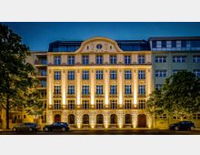 Beleuchtete historische Gebäudefassade in einer Stadtstraße bei Nacht, mit symmetrischen Fensterreihen, verziertem Mittelgiebel und einem rustizierten Sockel mit Rundbogenöffnungen. Vor dem Haus stehen Bäume und parkende Autos, rechts und links schließen moderne Nachbargebäude an.