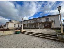 Helles, zweistöckiges Gebäude mit weißen Fassaden, großen Holzfenstern und einem Säuleneingang. Im Vordergrund sind breite Treppenstufen, ein gepflasterter Vorplatz sowie Laternen und ein Grünabfallbehälter zu sehen, umgeben von einer leicht bewachsenen Landschaft.