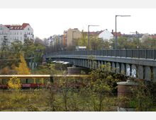 Straßenszene mit einer Brücke aus Stahl und Beton, die von Straßenlaternen gesäumt wird, und dahinter einer Reihe von mehrstöckigen Wohnhäusern. Im Vordergrund fährt eine rote S-Bahn zwischen Bäumen und Gestrüpp entlang.
