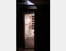Blick in einen dunklen, verlassenen Kellerraum mit einer offenstehenden Tür, die in einen weiteren, teils gefliesten Raum führt. An der Rückwand des zweiten Raums sind Rohrleitungen und technische Installationen sichtbar.