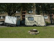 Wohnwagen und Anhänger stehen in einer Wohngegend vor einem mehrstöckigen Wohngebäude aus Beton mit brauner Fassade und weißen Fenstern. Im Vordergrund ist eine Wiese mit einem runden Betondeckel sichtbar.