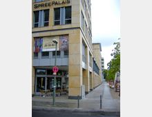 Ein modernes, helles Gebäude, bezeichnet als "Spree Palais", mit glatten hellgelben Fassaden und großen Fenstern. Im Erdgeschoss befinden sich ein Café mit Außensitzplätzen und ein Gehweg, der an Bäumen und weiteren Gebäuden entlangführt.