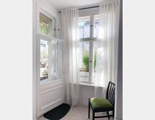 Helle Ecke eines Wohnraums mit großen weißen Holzfenstern, transparenten Vorhängen und Blick auf grüne Pflanzen und Gebäude im Außenbereich. Links eine geöffnete Balkontür, rechts ein Stuhl mit grünem Sitzpolster.