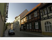 Schmale, gepflasterte Straße mit Fachwerkhäusern auf der rechten Seite, darunter ein Hotel mit einem Schild. Links befindet sich ein modernes Gebäudeschild mit der Aufschrift "Hotel". Im Hintergrund weitere Gebäude und Bäume bei sonnigem Wetter.