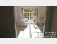 Heller Wohnraum mit großen bodentiefen Sprossenfenstern, durch die Sonnenlicht einfällt. Der Raum ist mit einem weißen Sessel, einem kleinen Tisch mit Stühlen und rosa Vorhängen ausgestattet, der Holzboden verstärkt die helle und freundliche Atmosphäre.