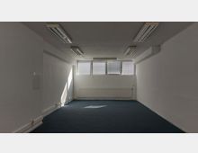 Ein leerer, rechteckiger Raum mit weißen Wänden, blauem Teppichboden und mehreren rechteckigen Fenstern in der oberen Wand am Ende des Raums. Die Decke ist mit rechteckigen Deckenleuchten und an der Wand ist ein langgezogener Heizkörper unter den Fenstern angebracht.