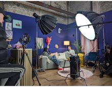 Studio-Set in einer hohen Industriehalle mit blau gestrichenen Wänden, grauem Sofa, Stehlampe, Vorhängen und dekorativen Bildern. Im Vordergrund stehen Kamera auf Stativ sowie große Softbox-Leuchten und weiteres Filmequipment auf Holzboden mit rundem Teppich und kleinem Tisch.