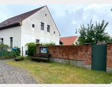 Weißes Wohnhaus mit Satteldach neben einer roten Backsteinmauer, in die ein grünes Holztor sowie ein Schaukasten integriert sind. Vor der Mauer befindet sich eine Holzbank, umgeben von einer kleinen Rasenfläche und einem gepflasterten Gehweg.