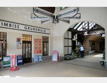 Eingangsbereich eines Bahnhofsgebäudes mit einem Imbiss („Imbiss Grunewald“) auf der linken Seite und einem Blumenstand auf der rechten Seite unter einem verglasten Dach. Die Architektur zeigt helle Wände, Holzverzierungen und Bögen, und auf dem Boden liegen beige Fliesen.