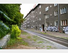 Straßenszene mit mehrstöckigen grauen Wohngebäuden auf der rechten Seite, teils mit Graffiti bedeckt. Links grenzt eine begrünte Fläche mit Büschen und Bäumen, daneben ein schmaler Gehweg.