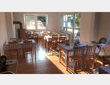 Ein heller Frühstücksraum mit mehreren Holztischen und Stühlen, die mit weißen Tischdecken gedeckt sind. Durch große Fenster fällt Sonnenlicht herein, und im Hintergrund ist eine Außenterrasse sichtbar.