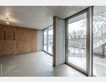 Minimalistisch gestalteter Innenraum mit Betonboden, einer Wand aus Sperrholz und großen Glasfenstern, die auf eine Terrasse mit Außentreppe und Blick auf ein Nachbargebäude führen.