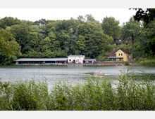 Ein See mit Schilf im Vordergrund, dahinter eine Bootsanlegestelle mit überdachten Ruderbooten und einem weißen Gebäude. Im Hintergrund steht ein gelbes Haus, umgeben von dichtem Wald.
