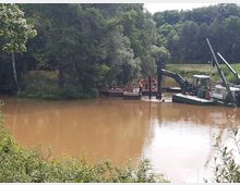 Ein braun gefärbter Fluss oder Teich, umgeben von Waldgebiet, mit Maschinen und Arbeitern auf schwimmenden Plattformen, die offenbar Arbeiten im Wasser durchführen. Im Hintergrund sind Bäume und dichtes Grün zu sehen.