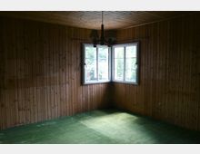 Kleiner Raum mit vertäfelten Holzwänden und -decke, grünem Teppichboden und einem Doppelfenster mit Blick ins Freie. An der Decke hängt eine schlichte Lampe mit drei Schirmen.