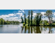Flusslandschaft mit ruhiger Wasseroberfläche, umgeben von grünen Bäumen und Vegetation; rechts ein kleines reetgedecktes Gebäude. Der Himmel ist leicht bewölkt mit blauem Himmel im Hintergrund.