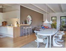 Moderne Wohnküche und Essbereich mit hellem Holzboden und minimalistischer Einrichtung. Linke Seite zeigt eine weiße Küchenzeile mit Holzarbeitsfläche, rechts ein Essbereich mit weißem Tisch, geschwungenen Stühlen und großen Hängeleuchten; im Hintergrund befindet sich eine Sitzecke mit grauem Sofa und Pflanzen.