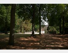 Parklandschaft mit hohen Bäumen, deren Blätter Schatten auf den Boden werfen, im Hintergrund eine klassizistische Steinbalustrade und ein Gebäude mit Säulen. Der Weg führt durch die Szenerie, gesäumt von schattigen Bereichen und Lichtungen.