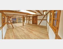 Innenansicht eines Fachwerkraums mit weiß verputzten Wänden, sichtbaren Holzbalken und einem Holzboden. Im hinteren Bereich steht ein Esstisch mit Stühlen, während eine Holztür und Fenster mit Holzrahmen die Wände prägen.