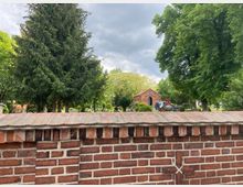 Eine Backsteinmauer im Vordergrund trennt den Blick auf eine grüne Parklandschaft mit hohen Nadel- und Laubbäumen. Im Hintergrund steht ein kleines Backsteingebäude mit Spitzbogenfenster, umgeben von weiteren Bäumen und vereinzelten Fahrzeugen.