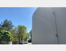 Außenbereich mit einem großen, runden, weißen Gebäude rechts im Bild und grünen Bäumen auf der linken Seite. Im Hintergrund sind ein Parkplatz mit parkenden Autos und ein klarer blauer Himmel zu sehen.