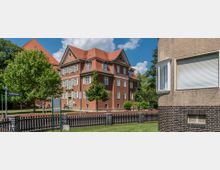 Wohngebiet mit historischen Gebäuden: Ein dreistöckiges, rot verputztes Gebäude mit spitzem, rotem Dach und weißen Sprossenfenstern, umgeben von Bäumen, einem Zaun und gepflegtem Grün. Rechts im Vordergrund ist ein moderner Wohnbereich mit einer abgerundeten Fensterecke und schwarzer Backsteinfassade zu sehen.