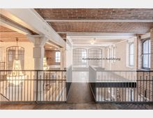 Innenraum eines industriell gestalteten Gebäudes mit Backsteinwänden, freiliegender Ziegeldecke und großen Fenstern. Zu sehen ist eine Galerie mit Geländern, Kronleuchtern im unteren Bereich und einem Bereich, der als „Konferenzraum in Ausführung“ gekennzeichnet ist.