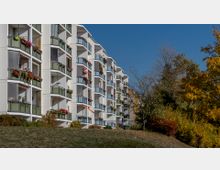 Mehrstöckiges Wohngebäude mit weißen Fassaden und vielen Balkonen, die mit Blumen geschmückt sind. Im Vordergrund eine Rasenfläche, im Hintergrund Herbstbäume mit gelbem und rotem Laub.