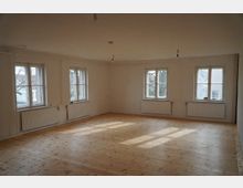 Heller, leerer Raum mit Holzboden, weißen Wänden und vier Fenstern, durch die Tageslicht einfällt. Die Fenster sind mit Heizkörpern darunter ausgestattet, und die Decke hat mehrere unbeleuchtete Lampenfassungen.