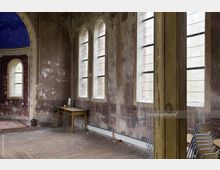 Innenraum eines verfallenen Gebäudes mit hohen, schmalen Bogenfenstern, vergilbten Wänden und abgenutztem Ziegelboden. Ein Holztisch und gestapelte Stühle stehen an der Seite, während ein Teil einer blauen, verzierten Decke sichtbar ist.