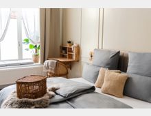 Gemütliches Schlafzimmer mit einem Bett, das graue Bettwäsche und beigestes Dekokissen hat. Im Hintergrund ein Fenster mit hellen Vorhängen, eine Holzschreibtischecke mit Pflanzen und ein geflochtener Rattankorb auf dem Bett.