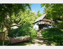 Ein kleines, weißes Landhaus mit grauem Satteldach steht inmitten einer grünen, waldreichen Umgebung. Im Vordergrund befindet sich eine einfache Liege unter einem Baum, umgeben von hohen Gräsern und Schatten.