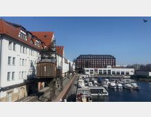 Hafenansicht mit weißen Gebäuden und rotem Ziegeldach auf der linken Seite, einem historischen Hafenkran in der Mitte und einem modernen, mehrstöckigen Ziegelgebäude im Hintergrund. Im Vordergrund liegen mehrere Boote am Wasseranleger vor Anker.