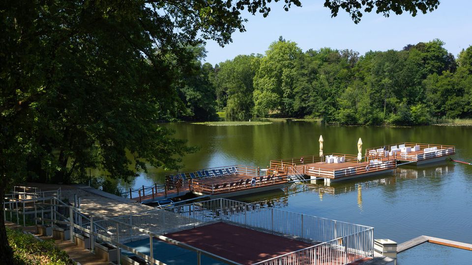 Lakeside recreation area with a calm green lake bordered by dense trees, and several large wooden floating docks with railings, seating, and sun umbrellas. In the foreground, stepped concrete terraces and metal railings lead down to the water.