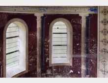 Innenansicht eines historischen Gebäudes mit zwei schmalen, bogenförmigen Fenstern und dekorativen Säulen an einer dunkelrot gestrichenen Wand. Die Wand zeigt Spuren von Verwitterung und zahlreiche eingeritzte Graffiti, daneben sind ornamentale Fliesen sichtbar.