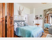 Schlafzimmer mit hellem, stilvollem Interieur. Ein Bett mit einem schwarz-weiß gemusterten Kopfteil und blauen Akzenten steht im Mittelpunkt, darüber hängen gerahmte Bilder und eine dekorative Tierkopf-Skulptur; daneben befinden sich ein großer verzierter Spiegel und eine Kommode mit Lampe.