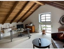 Ein Arbeitszimmer unter einer schrägen Holzdecke mit sichtbaren Balken. Der Raum enthält einen Glastisch mit Computer, ein Regal, eine Couch, einen runden Beistelltisch, eine Zimmerpflanze vor einem Fenster mit Sprossen und weiteren Möbeln in warmen Holztönen.