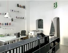 Helles Büro mit weißen Wänden, einem Schreibtisch, auf dem Arbeitsutensilien stehen, schwarzen Regalen mit Aktenordnern und Modellfiguren auf Wandregalen. Im Hintergrund befinden sich ein Kicker und ein Modellsegelboot, darüber ein grünes "B"-Logo an der Wand.