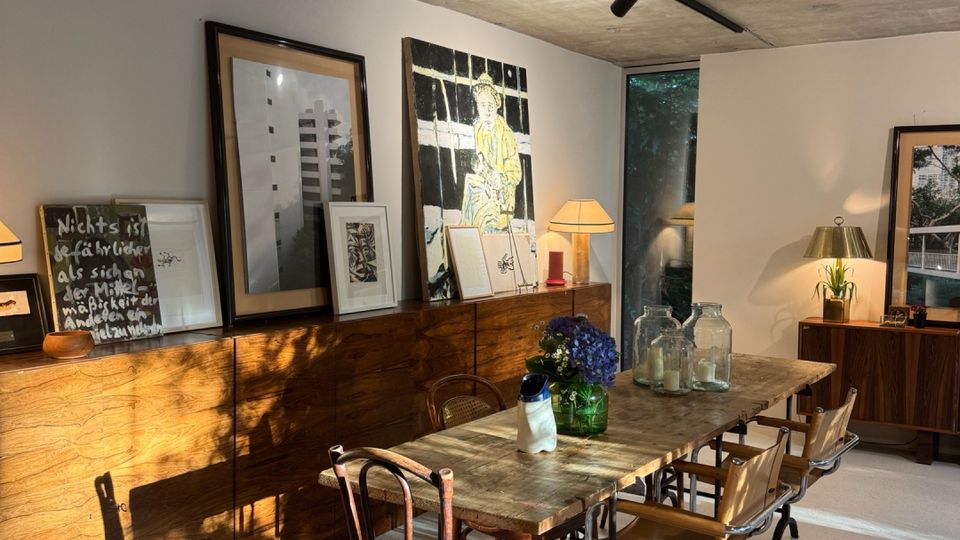 Dining area in a modern, minimalist home with a raw concrete ceiling, a long wooden dining table, and mixed chairs. A low wooden sideboard displays framed artwork and table lamps, with sunlight casting long shadows across the light floor near a tall window.
