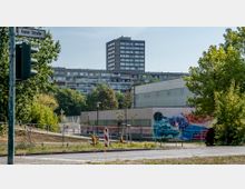 Städtische Szene mit einer großen grauen Wohnanlage im Hintergrund und einer flachen Sport- oder Lagerhalle im Vordergrund, deren Seitenwand mit buntem Graffiti verziert ist. Im Vordergrund sind eine Straße mit einem Straßenschild "Kieler Straße", eine Ampel sowie Bäume und Grünflächen zu sehen.