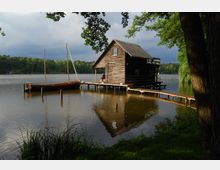 Holzhaus auf Stelzen am Ufer eines Sees mit einem Steg und einem Segelboot. Im Hintergrund Wald und bewölkter Himmel, im Vordergrund Gras und Bäume.