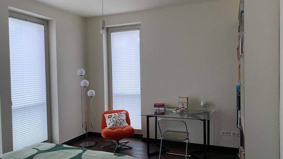 Helles Schlafzimmer mit zwei hohen Fenstern mit Jalousien, dunklem Holzboden und weißen Wänden. Im Raum stehen ein Bett mit gemusterter Decke, ein orangefarbener Sessel, eine Stehlampe mit mehreren Kugelleuchten sowie ein gläserner Schreibtisch mit klappbarem Stuhl und einem hohen Regal an der Wand.