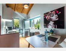 Kleine, moderne Küche mit Holzboden und einer hohen Decke mit sichtbaren Holzbalken. Rechts steht ein Tisch mit Dekorationsobjekten, während links eine kleine Spüle und ein Kühlschrank zu sehen sind; zwei Fenster bieten Blick ins Grüne.