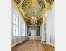 Barocker Innenraum mit dekorativem Holzparkett, weiß-goldenen Wänden und einer reich verzierten Decke mit Malereien und Ornamenten. Im Zentrum hängt ein prunkvoller Kronleuchter, und an der Stirnseite befindet sich eine Holztür mit gemalter Blumen-Dekoration, daneben ein dekorativer weißer Kachelofen.