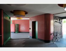 Überdachter Durchgang mit roten Wandfliesen und grünen Türen, dekoriert mit gelben Deckenleuchten. Im Hintergrund ist ein Fahrrad an einem kunstvoll verzierten Metalltor angelehnt.