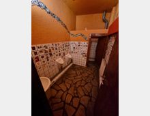 Öffentliche Toilettenanlage mit ungewöhnlichem Design: Wände aus orangefarbenem Putz und Dekorfliesen mit Mosaiken und Abbildungen, ein Boden aus unregelmäßigem Steinmuster, zwei Urinale an einer Kachelwand und ein Waschbecken links. Ein Metallrohr verläuft an der Decke.